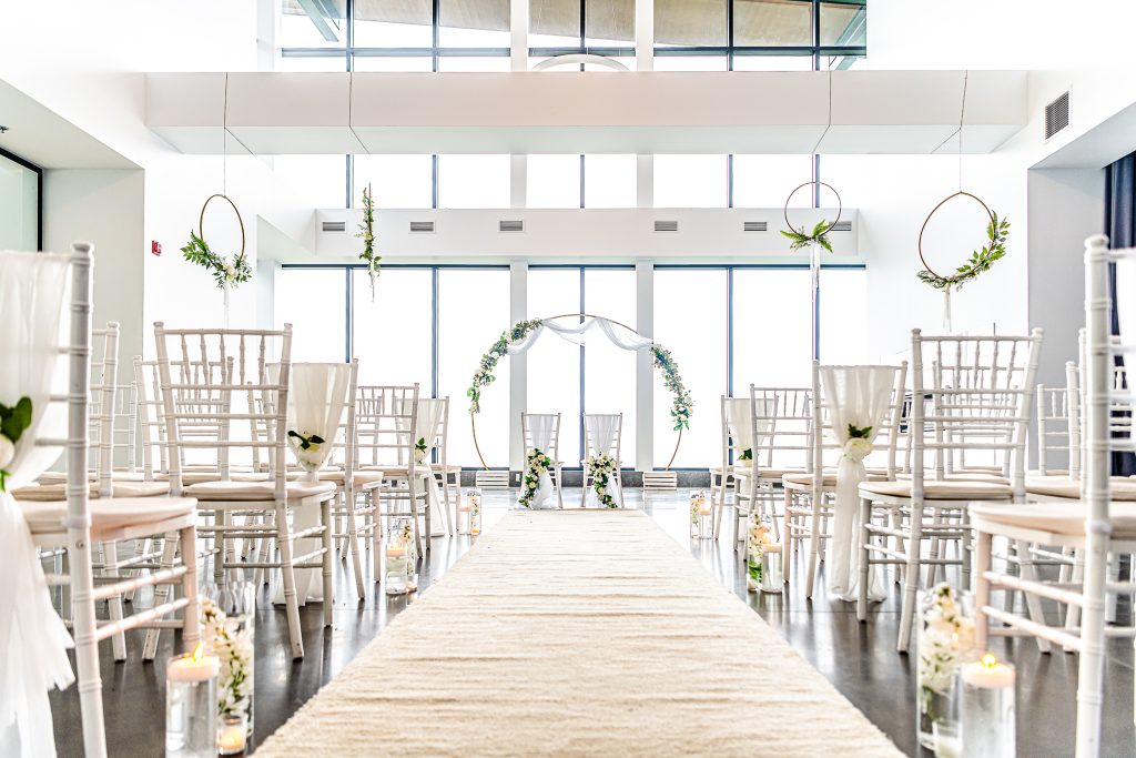 Salle de mariage lumineuse avec chaises blanches, un tapis beige, et un autel circulaire décoré de verdure, sous un plafond haut et moderne.