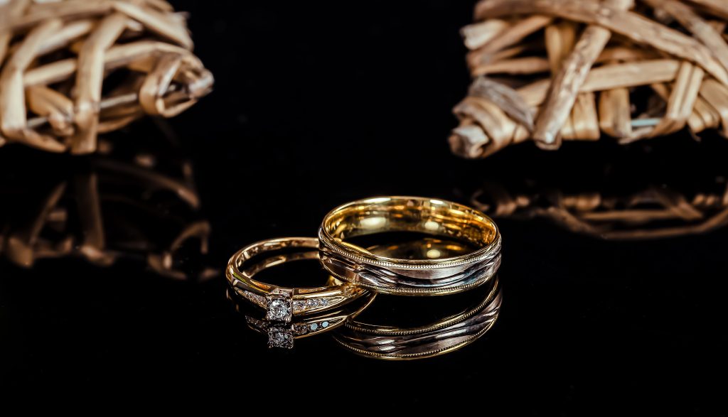 Deux alliances de mariage sur fond noir, comprenant une bague en or avec un diamant, encadrée par une autre en or lisse et une en argent avec des détails finement ciselés."