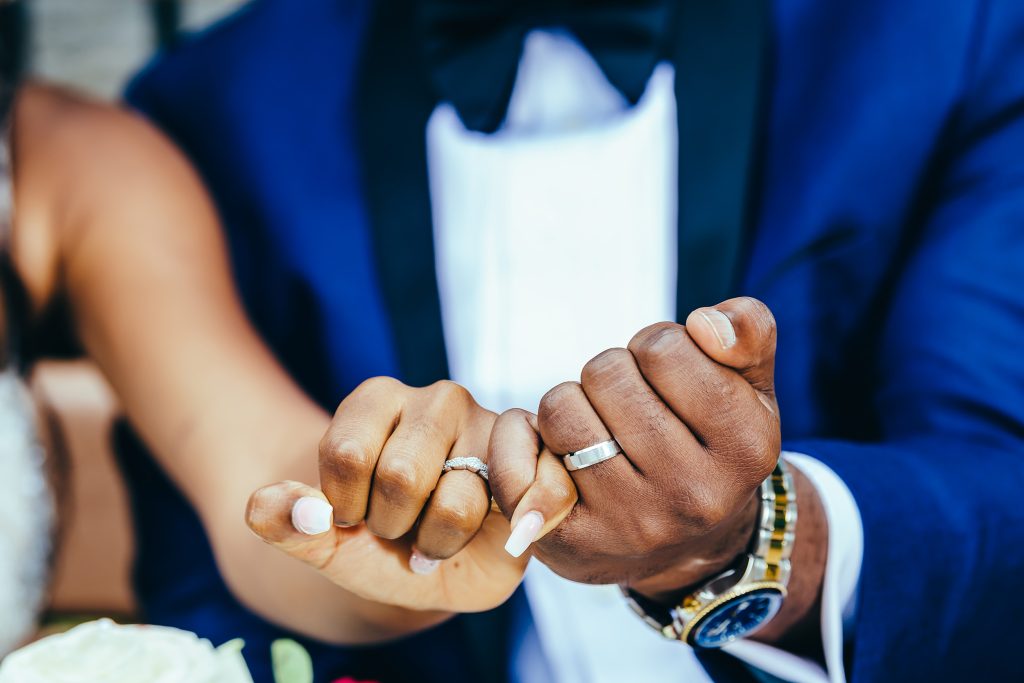 Mains des mariés joignant leurs doigts, montrant leurs alliances, sur un fond de leurs tenues de mariage bleues.