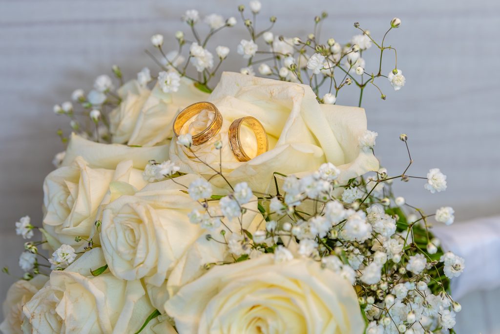 Alliances Dorées et Douceur Florale Deux alliances en or posées sur un bouquet de roses crème et de délicates fleurs blanches, représentant l'amour et la dévotion.
